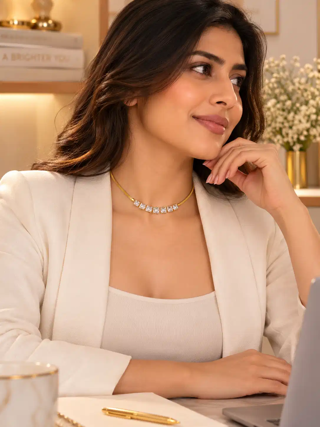 Professional woman wearing a gold-tone crystal necklace with a white blazer while seated at a laptop