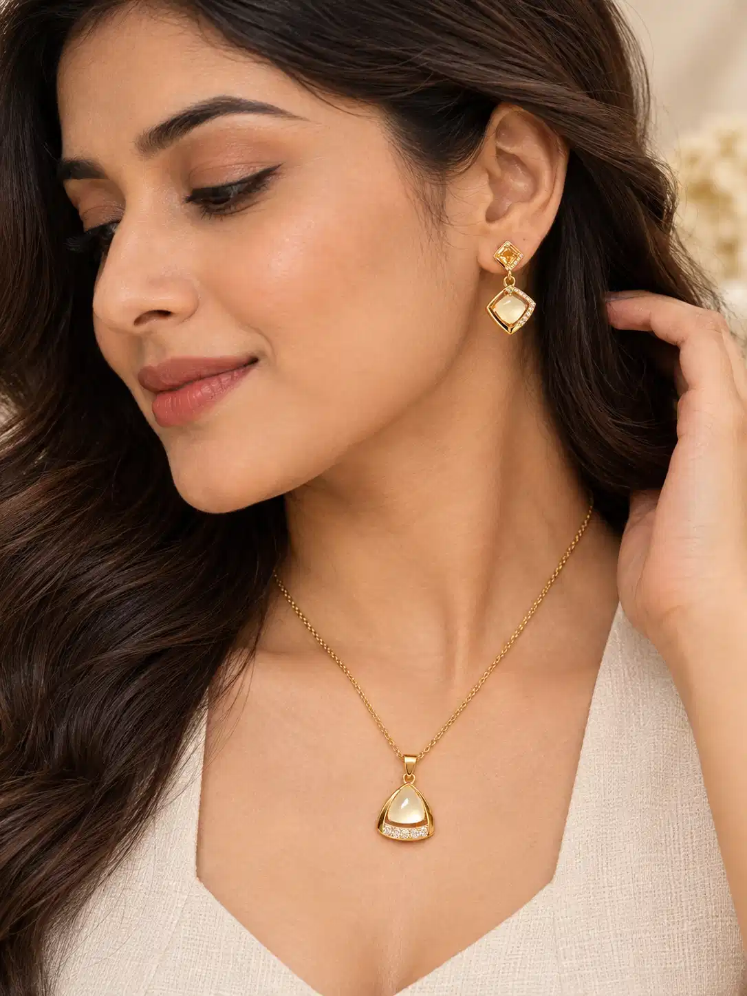 Woman wearing Ivora Glow gold pendant necklace and geometric drop earrings with an ivory outfit