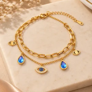 Gold-tone double-layer evil eye charm bracelet with crystal nazar centerpiece, blue teardrop charms and paperclip chain.