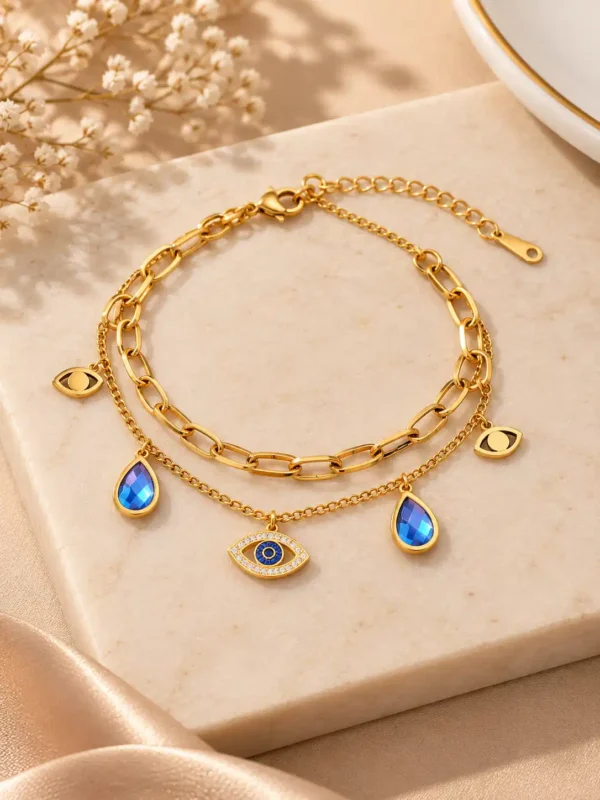 Gold-tone double-layer evil eye charm bracelet with crystal nazar centerpiece, blue teardrop charms and paperclip chain.