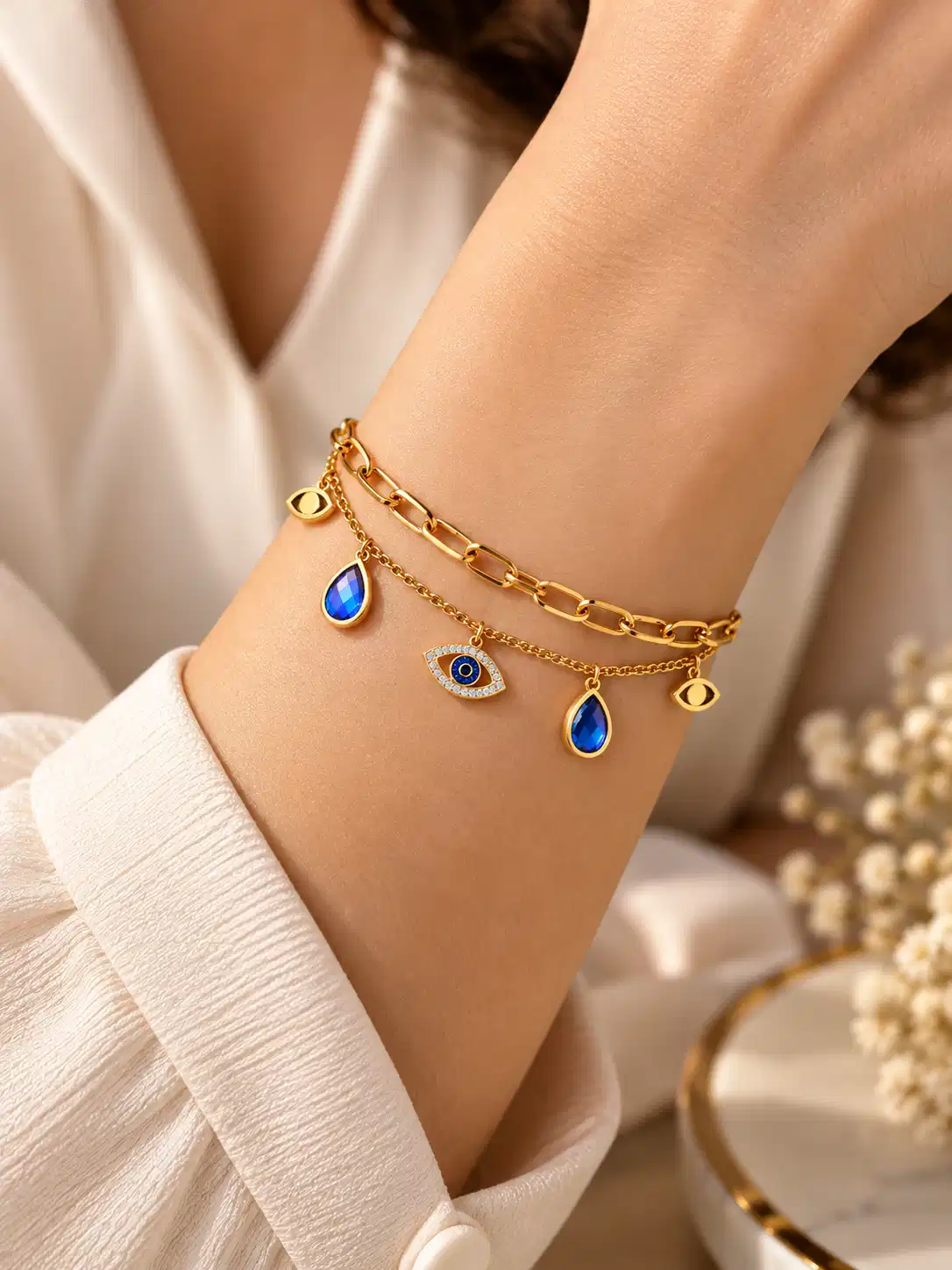 Woman wearing gold evil eye layered bracelet with blue charms on wrist, styled with a white outfit.
