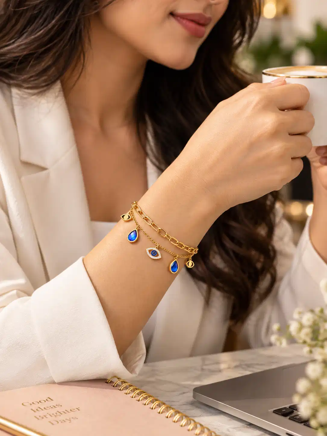 Woman wearing gold evil eye charm bracelet with blue drops at an office desk while dressed in a white blazer.